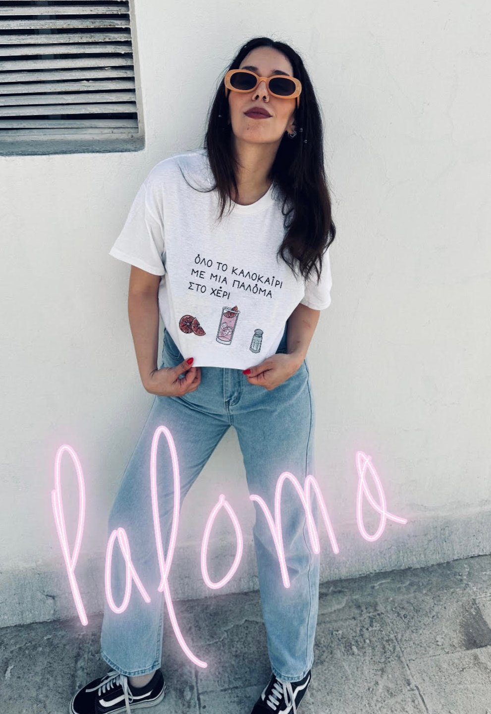 A girl wearing orange sunglasses standing and showing off her white unisex tee with the Greek text 'Όλο το καλοκαίρι με μια Παλόμα στο χέρι' on the chest, featuring below the text illustrations of a pink grapefruit, a paloma cocktail and a salt shaker
