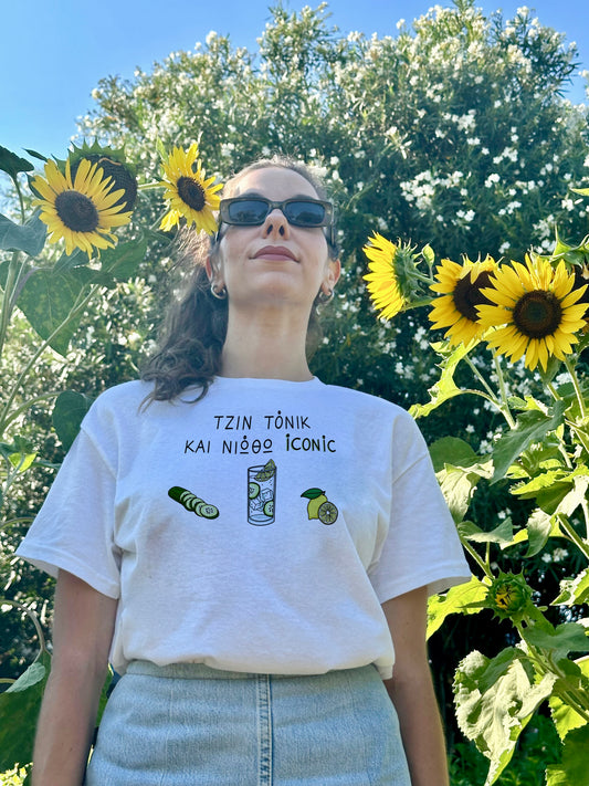 A girl standing among sunflowers, wearing sunglasses and a white unisex tee with the Greek text 'Gin Tonic και νιώθω iconic' on the chest, featuring illustrations of a cucumber, gin tonic, and lemon