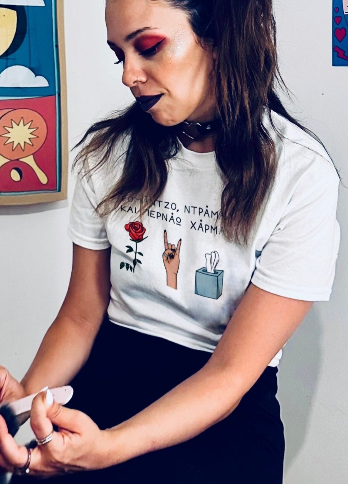 A girl sitting on a stool filing her nails wearing a white unisex tee with the Greek text 'Ρομάντζο, ντράμα και περνάω χάρμα' on the chest, featuring below the text illustrations of a red rose, a hand making the sign of the horns, and a packet of tissues