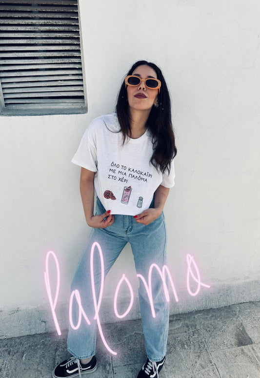 A girl wearing orange sunglasses standing and showing off her white unisex tee with the Greek text 'Όλο το καλοκαίρι με μια Παλόμα στο χέρι' on the chest, featuring below the text illustrations of a pink grapefruit, a paloma cocktail and a salt shaker