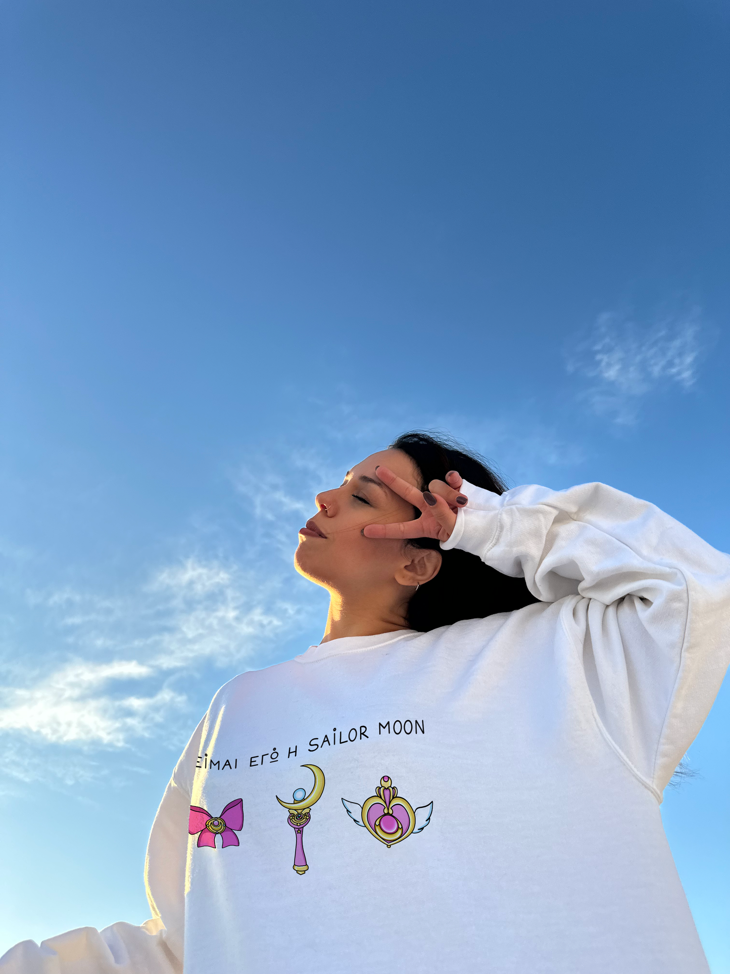 A girl with closed eyes making the v sign with her hand near her eyes, wearing a white sweatshirt featuring a Greek text on the chest 'Ειμαι εγώ η Sailor Moon'. Below the text is an illustration of Sailor Moon's signature items in pink tones: a ribbon, moon stick, and a heart.
